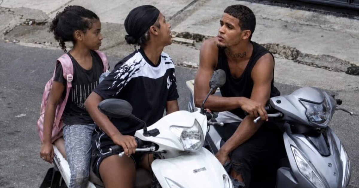 Three young people sit on two scooters in the street, chatting with each other; a girl with a pink backpack sits on the left.