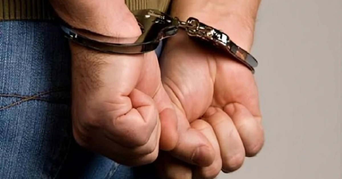 Person’s wrists are secured in shiny metal handcuffs behind their back, with blue jeans visible.