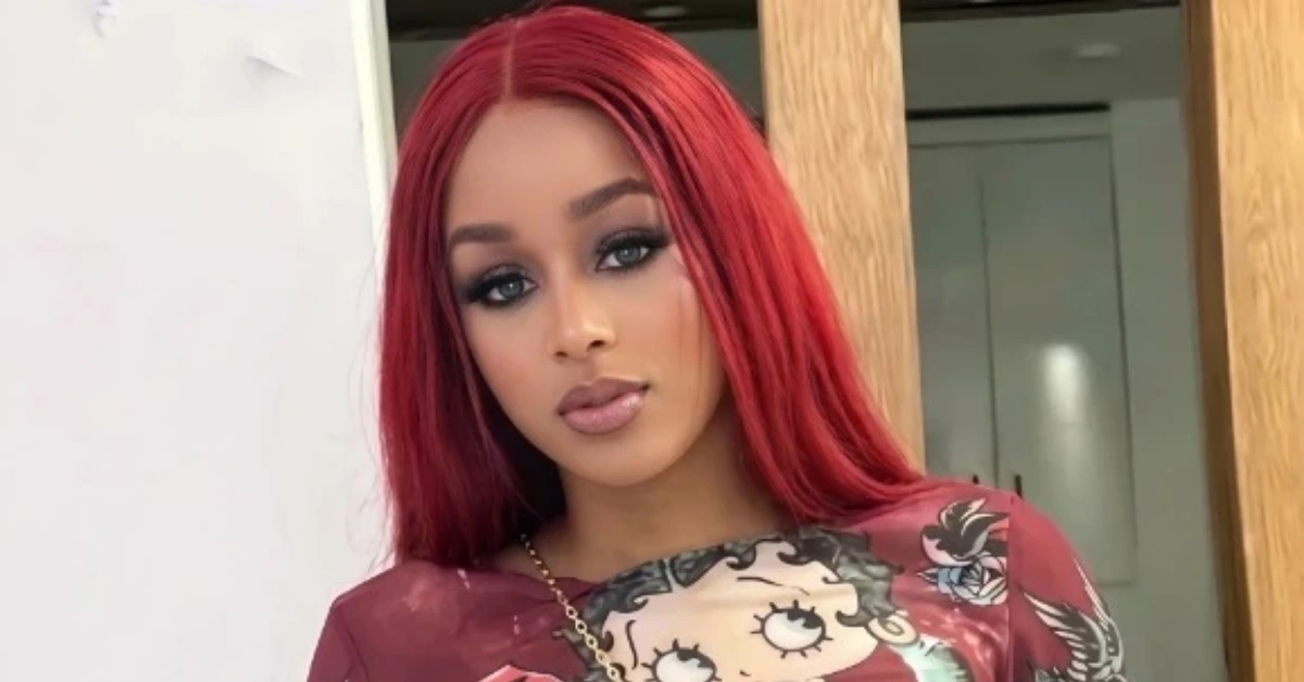 Young woman with long straight red hair and bold makeup poses near a doorway, wearing a burgundy shirt with a cartoon print.