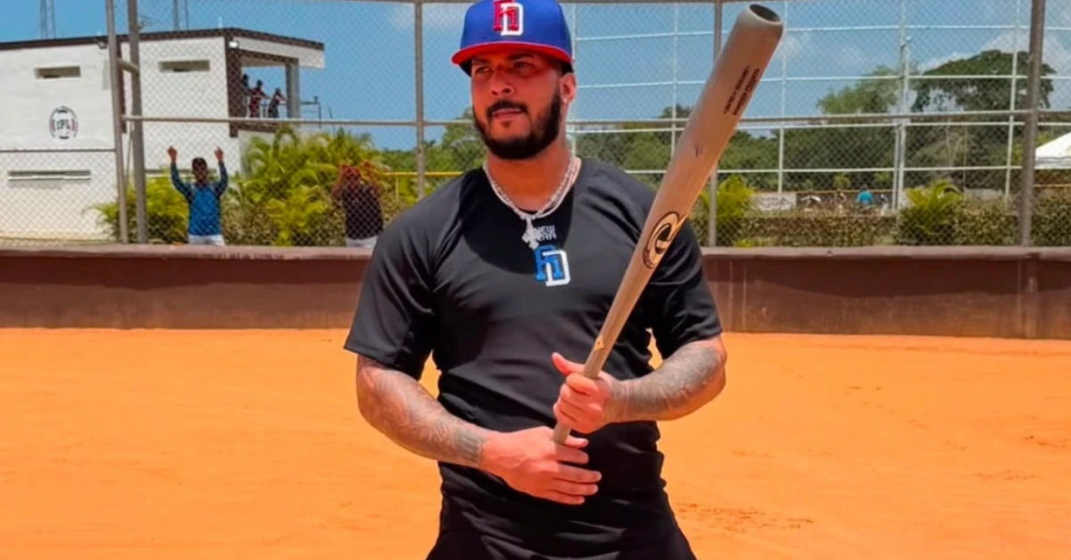Baseball batter standing on the dirt field, gripping a wooden bat and wearing a black shirt and blue-red cap, ready to swing.