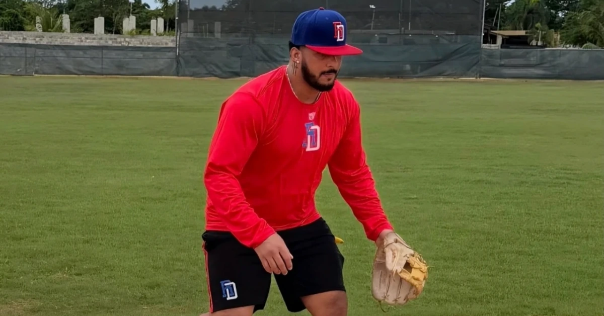 Baseball player in a red long-sleeve shirt and blue cap crouches with a glove, ready to field on a green outfield.
