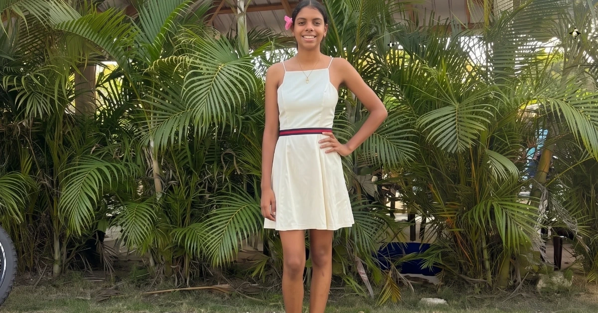 Girl in a light cream dress with a pink flower in her hair, standing with one hand on her hip among tropical plants.