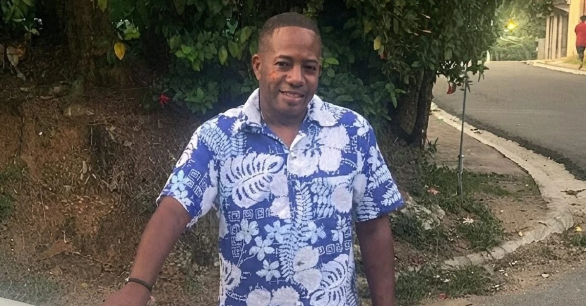 Smiling man in a blue and white tropical shirt standing on a sidewalk with trees behind him.