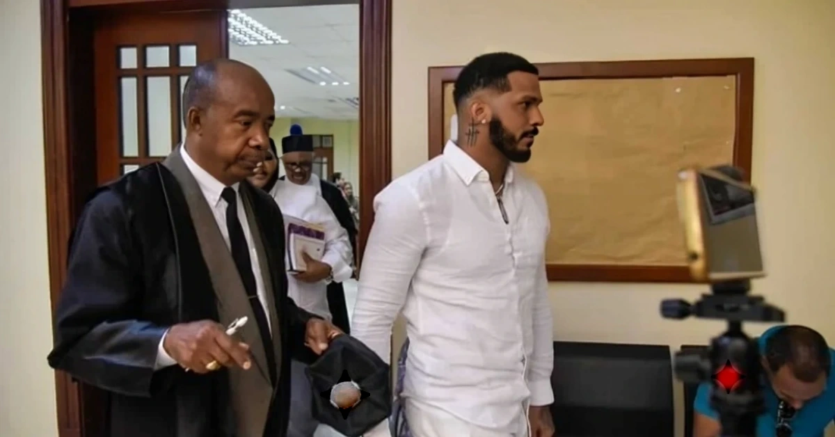Two men walk through a doorway in a hallway; one in a black judicial robe holding a hat, the other in a white shirt, as a camera on a tripod records nearby.