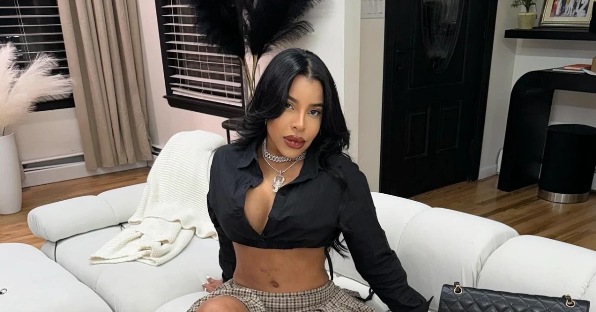 Woman with long black hair wearing a black cropped blouse and plaid skirt sits on a white sofa indoors.