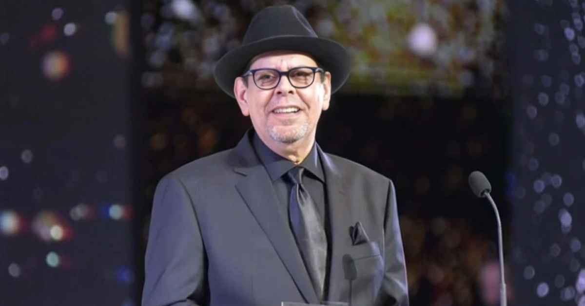 Man in a dark suit and tie wearing a fedora, standing at a podium with a microphone during a formal event.
