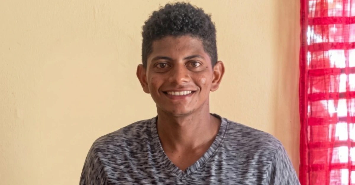 Smiling young man with short curly hair in a gray patterned shirt, standing indoors near a beige wall and red curtains.
