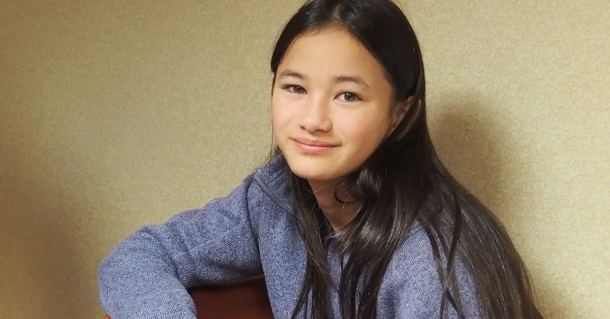 Portrait of a smiling young Asian woman with long black hair wearing a blue sweater, posing with a guitar beside her.