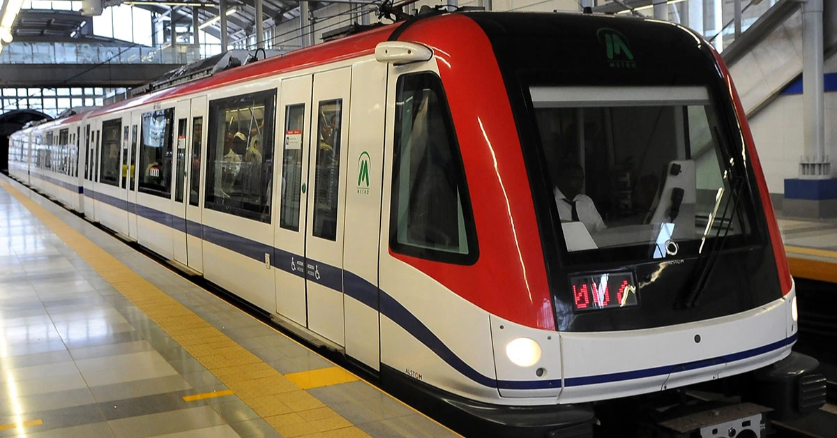 Modern white metro train with a red front and blue stripe at an indoor station platform, passengers visible through the windows.