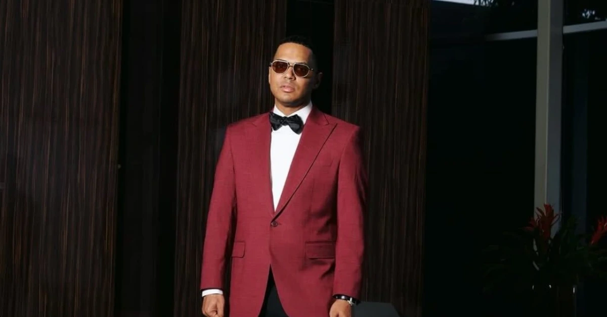 Man in a burgundy blazer with white shirt and black bow tie, wearing sunglasses, standing against a dark backdrop indoors.