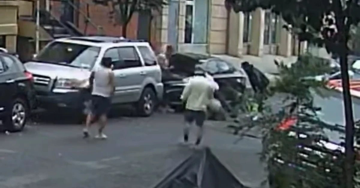 People stand and walk near a white SUV parked on a city street parking lane, with buildings in the background.