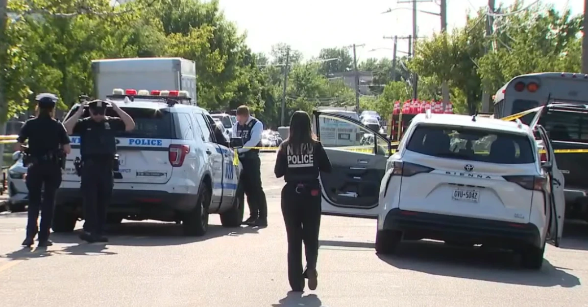 Police officers secure a street scene behind yellow caution tape, with patrol cars in the background and an open car door nearby.