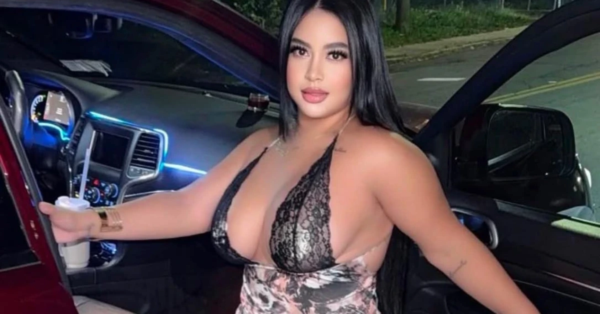 Woman wearing a black lace halter top leaning into a red car, holding a drink with a straw and a blue-lit dashboard in the background.