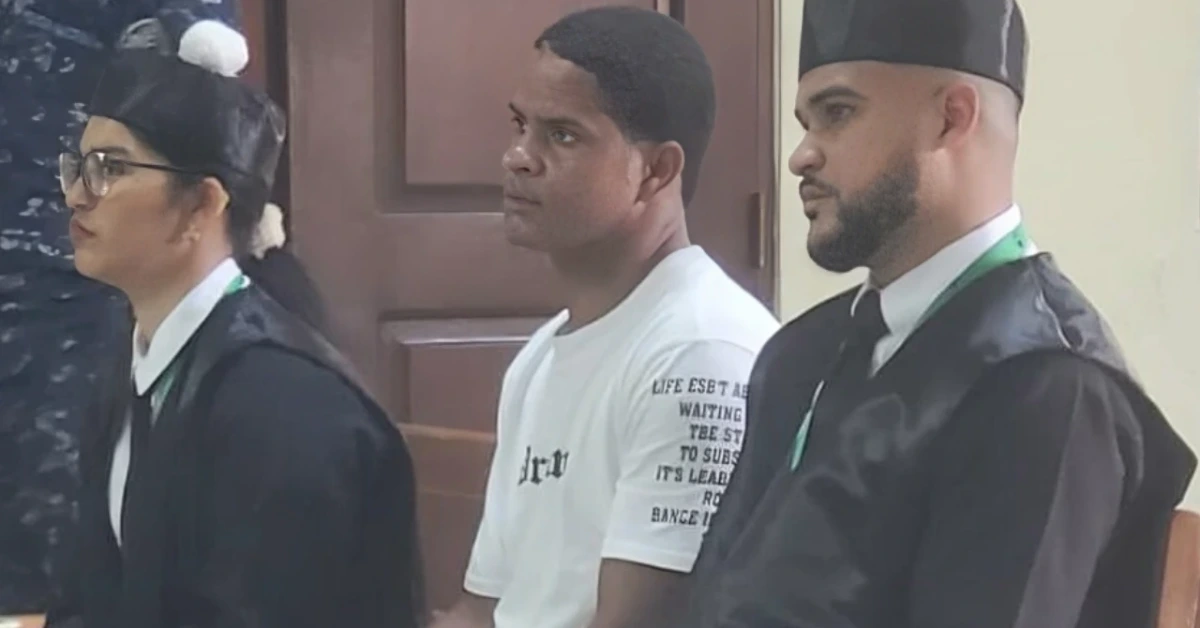 Three people sit in a row, two wearing black robes and graduation caps while a man in a white t-shirt sits between them, likely in a formal ceremony or court setting.