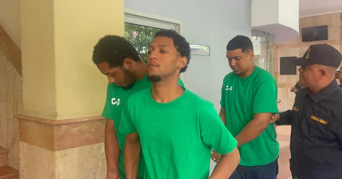 Three young men in green shirts walk in a building corridor, escorted by a uniformed security officer.