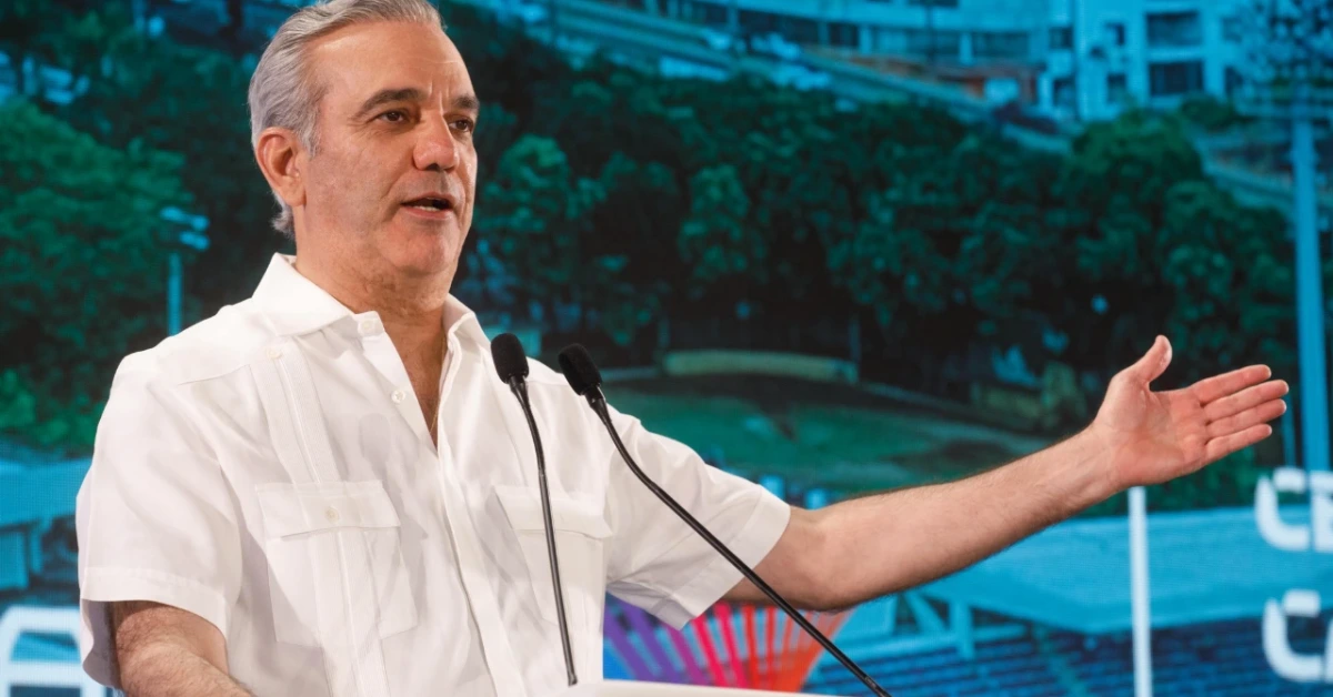 Man in a white short-sleeve shirt delivering a speech at a podium with two microphones, gesturing with an outstretched arm, against a blue-toned backdrop.