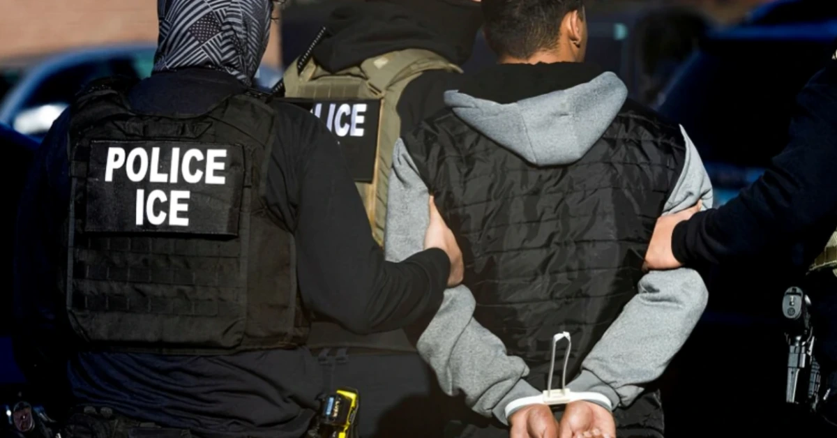 ICE police officers in tactical vests escort a handcuffed man in a gray hoodie from behind.