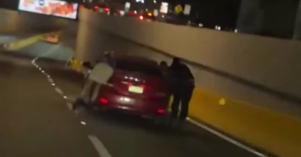 Red sedan pulled over on a dark highway at night with two people standing beside the trunk near a concrete barrier.