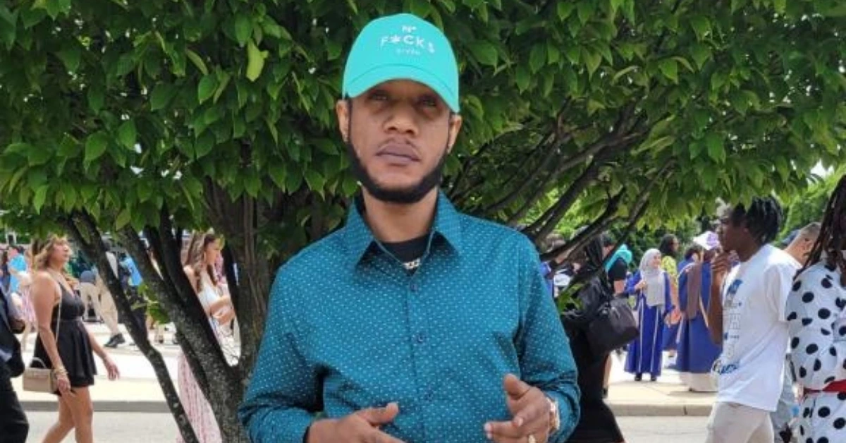 Man in a turquoise cap and dotted teal shirt stands before a leafy tree at an outdoor crowded event.