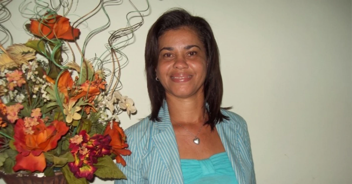 Smiling woman in a turquoise top and light blue striped blazer standing beside a large colorful flower bouquet.