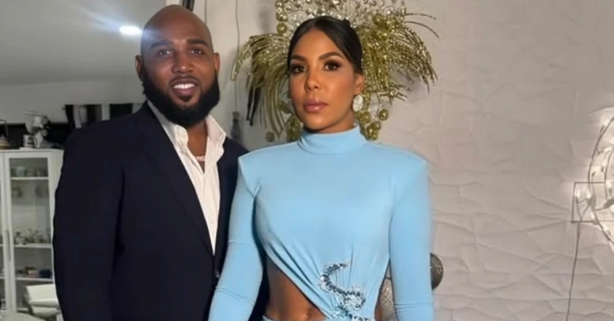 A man in a dark suit stands beside a woman in a light blue, high-neck dress with a side cutout and decorative embroidery in a stylish indoor setting.