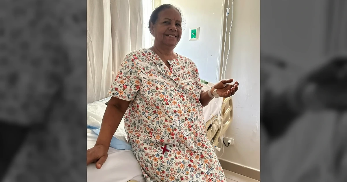 Pregnant woman in a floral hospital gown sitting on a hospital bed, smiling, with an IV line in her hand.