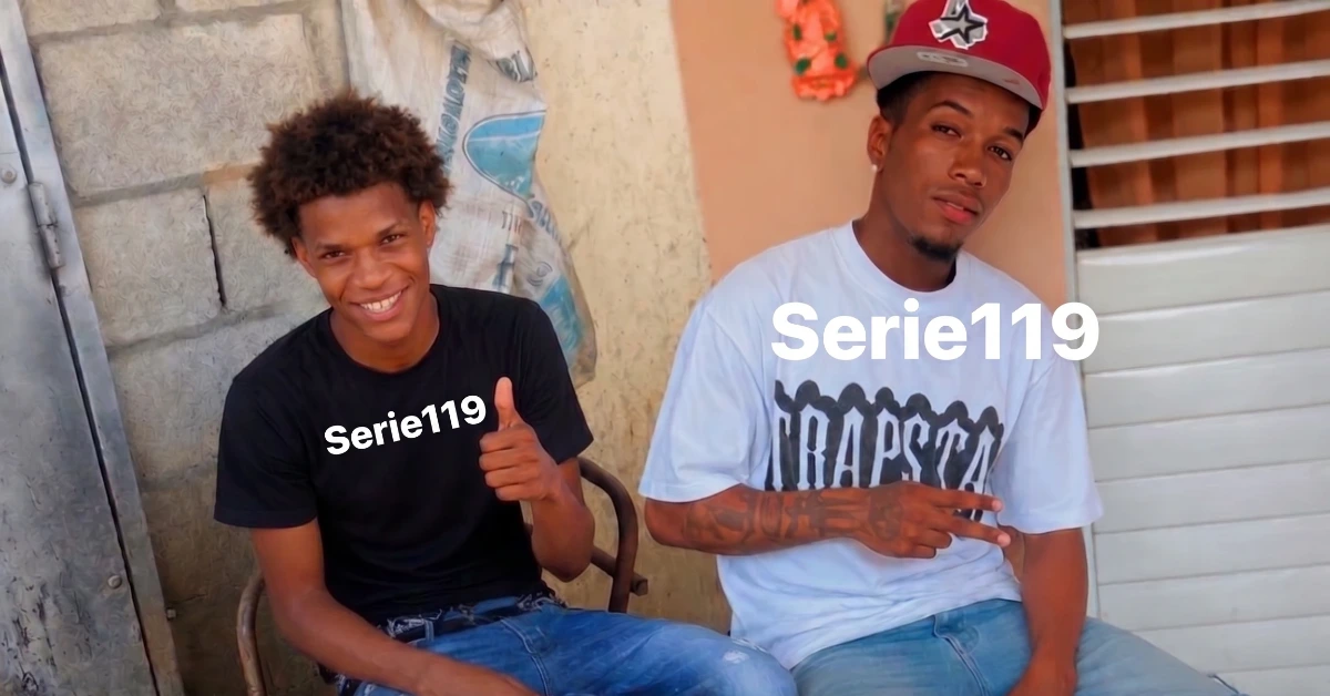 Two young men sit outdoors and smile at the camera; left man in black tee gives a thumbs‑up, right man in a white graphic tee rests with a hand gesture; 'Serie119' overlayed on their torsos.