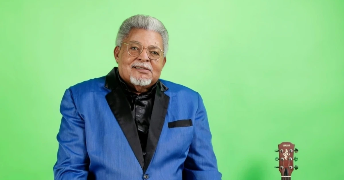 Portrait of an older man with white hair and glasses in a blue blazer, with a guitar visible on a bright green background.