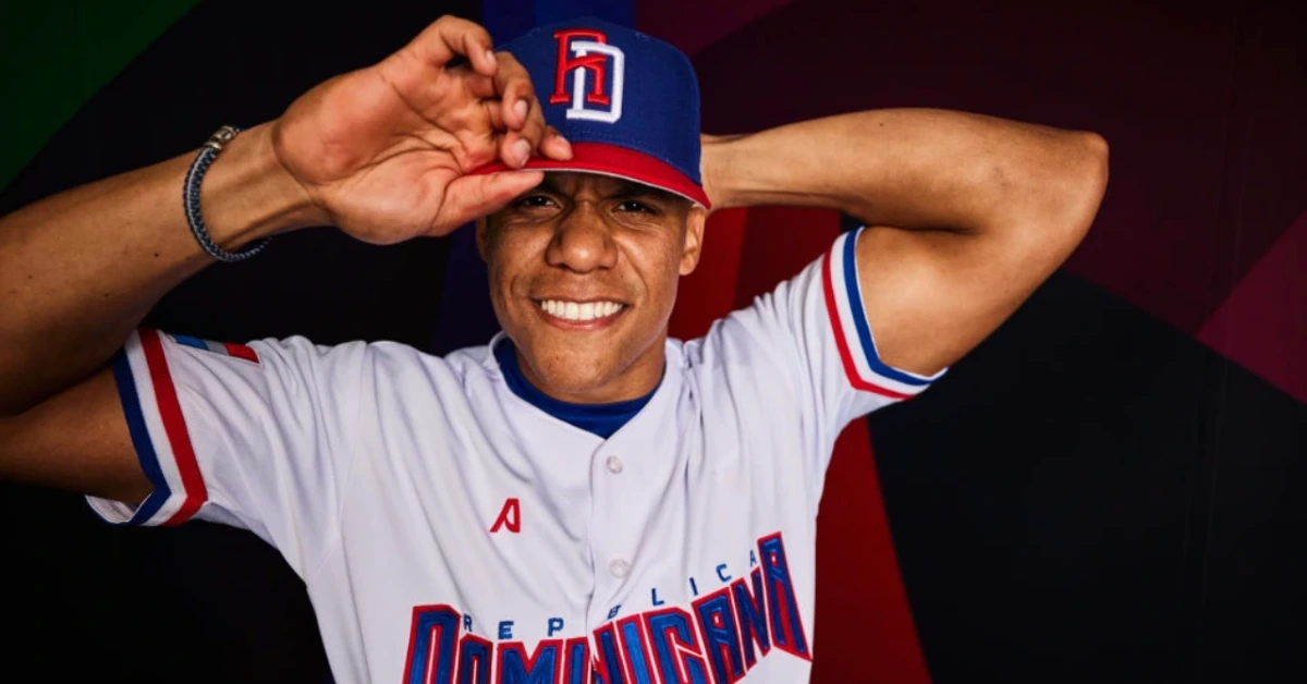 Smiling baseball player in white jersey with blue/red lettering and cap, saluting with one arm raised behind head.
