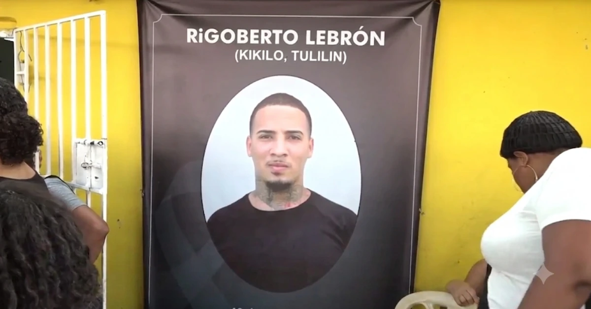 Memorial banner with a portrait of Rigoberto Lebrón (Kikilo, Tulilin) displayed on a yellow wall, people nearby viewing it