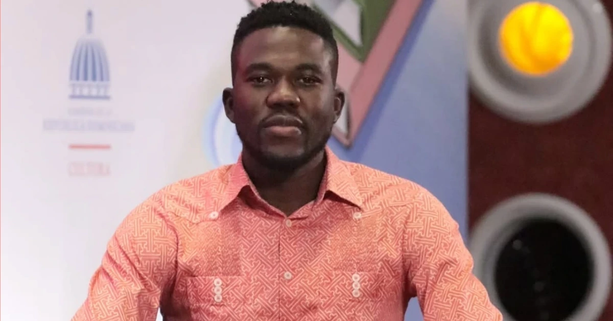 Portrait of a Black man with short hair and a trimmed beard wearing a pink patterned shirt, seated at a desk with a colorful backdrop behind him.