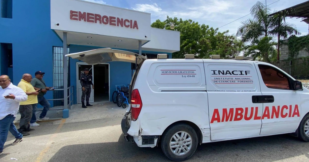 White ambulance with 'AMBULANCIA' in red letters parked at the blue hospital entrance labeled EMERGENCIA; several men stand nearby, and a security guard is at the doorway.