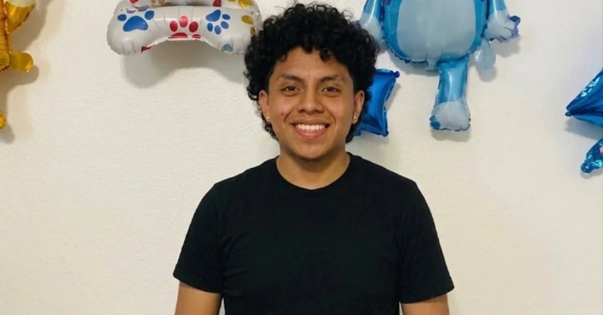Smiling person with short curly hair wearing a black t-shirt, standing in front of festive balloons in a party setting.