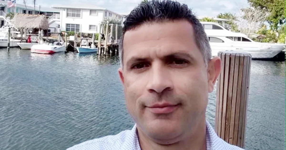 Portrait of a man taking a selfie by a marina with boats and houses in the background, calm water behind him.