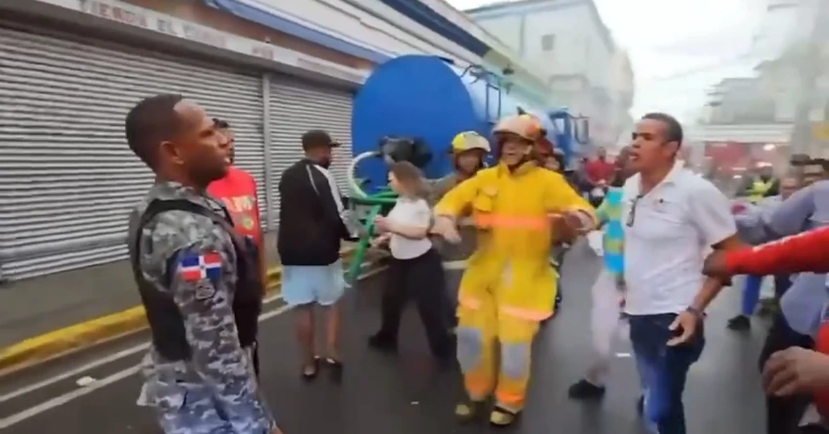 Emergency responders on a city street, a firefighter in yellow gear facing a military-style uniform amid smoke in the background.