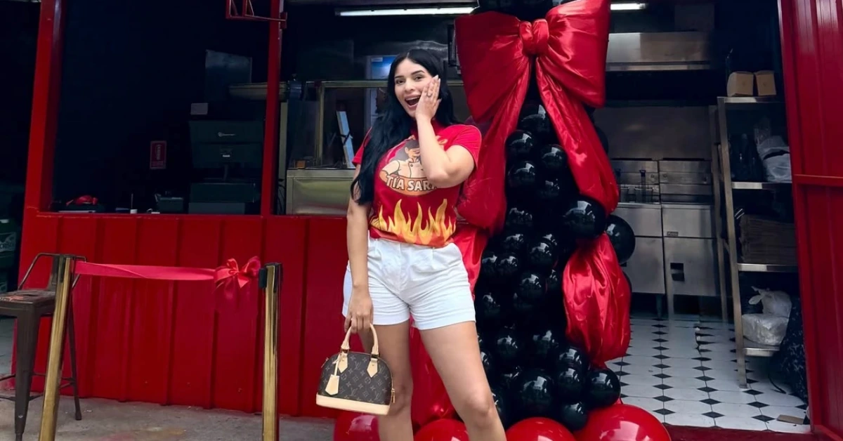 Woman in a red flame-themed shirt and white shorts poses at a storefront decorated with a large red bow and black balloon column.