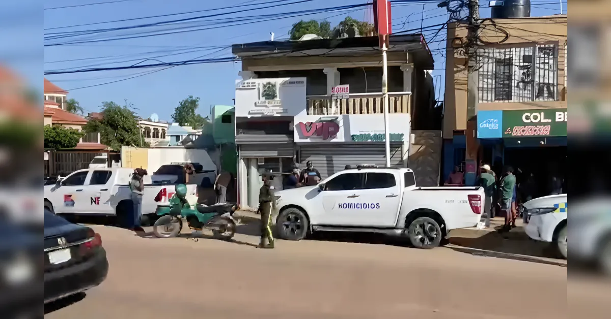 White police pickup with 'HOMICIDIOS' parked on a dusty street in front of small storefronts; officers and bystanders nearby, with motorcycles and other vehicles around.