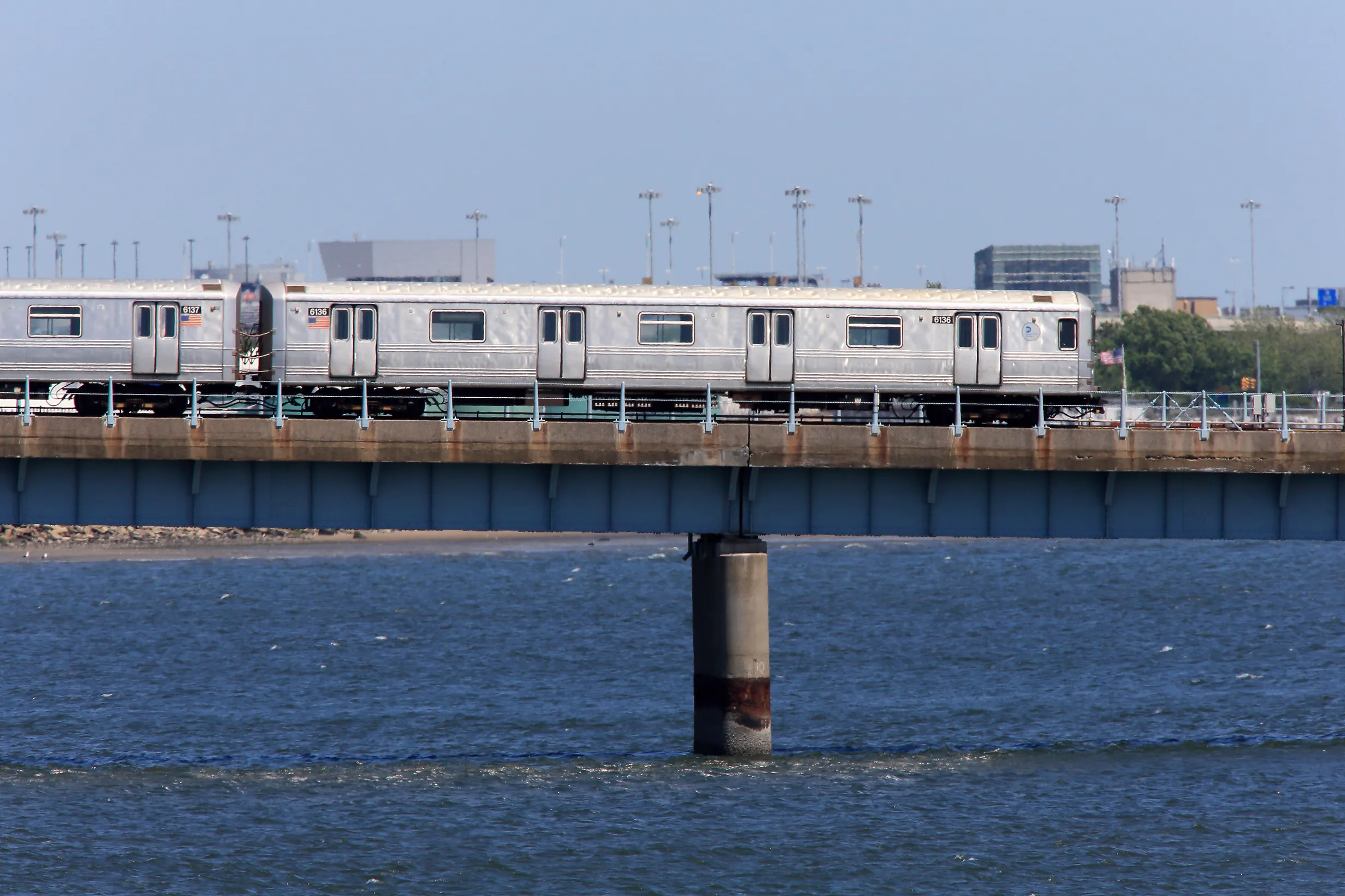 Silver commuter train cars on an elevated bridge spanning blue water, with city buildings in the distance under a clear sky.