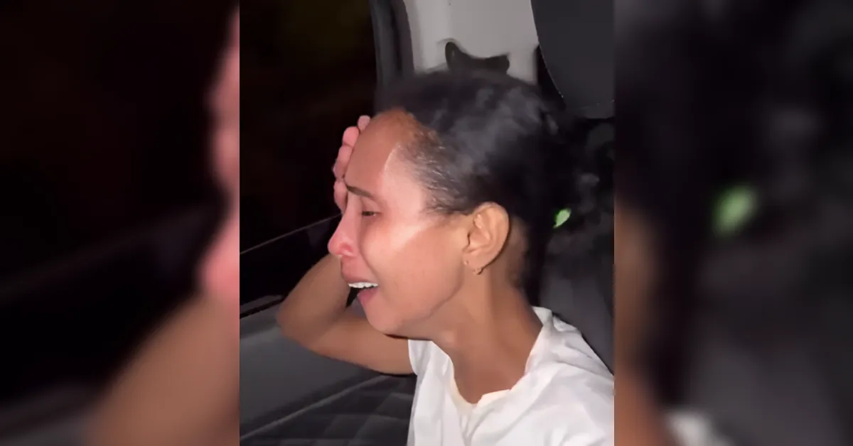 Woman in a white shirt sitting in a car at night, hand resting on her head while speaking