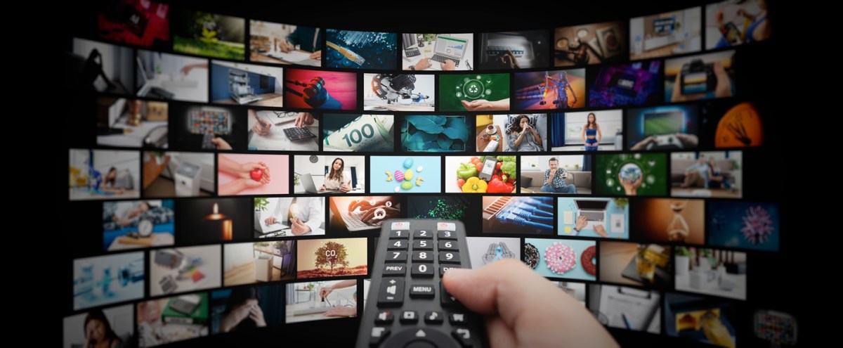 Hand holding a remote control in front of a curved wall of small screens showing assorted images and videos.