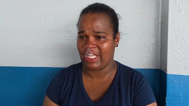 A woman in a navy shirt crying and speaking, tears on her face, standing against a gray wall with a blue lower panel.