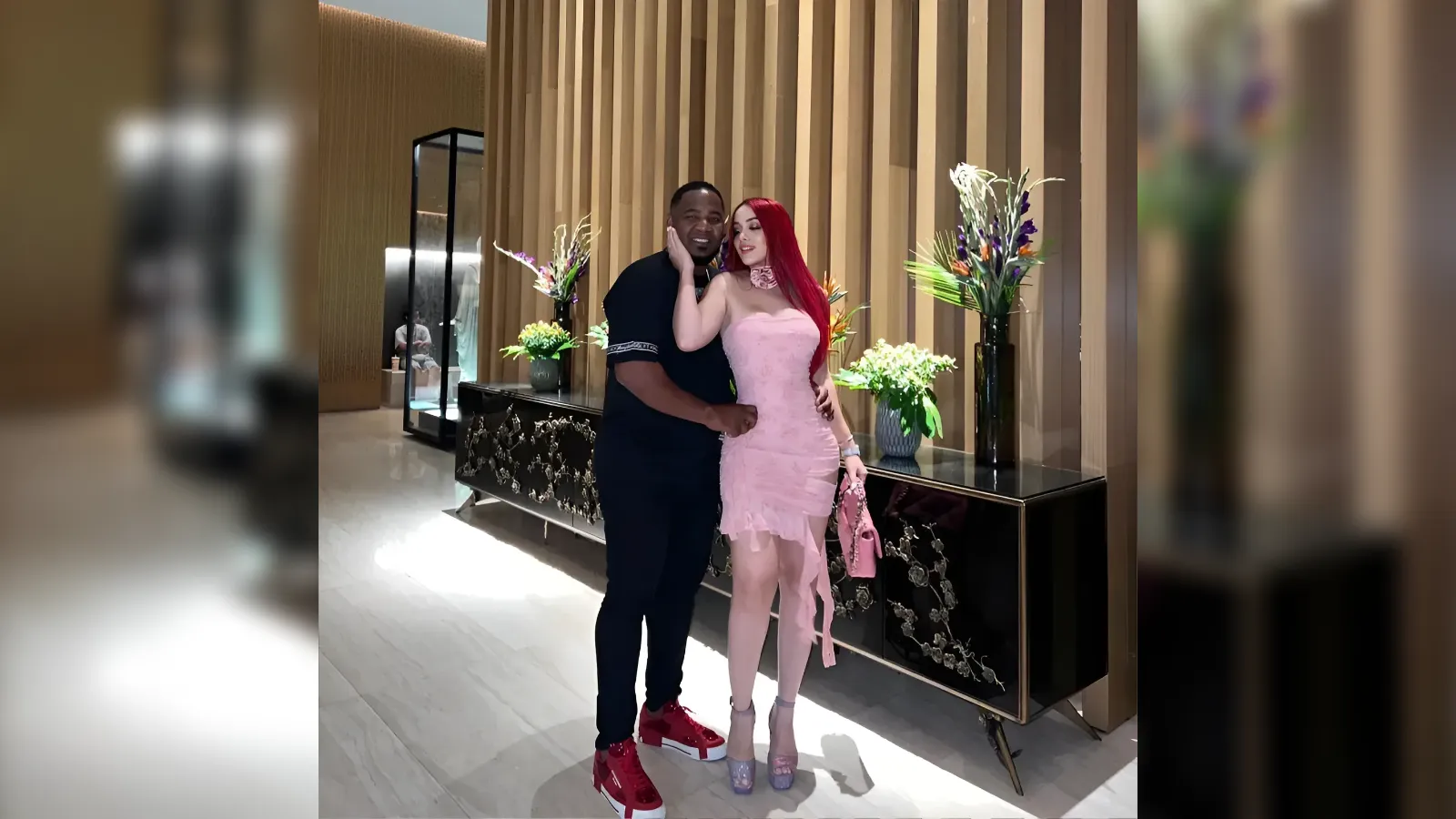 Couple posing together in a stylish lobby with a wooden paneled backdrop and flower arrangements behind them.