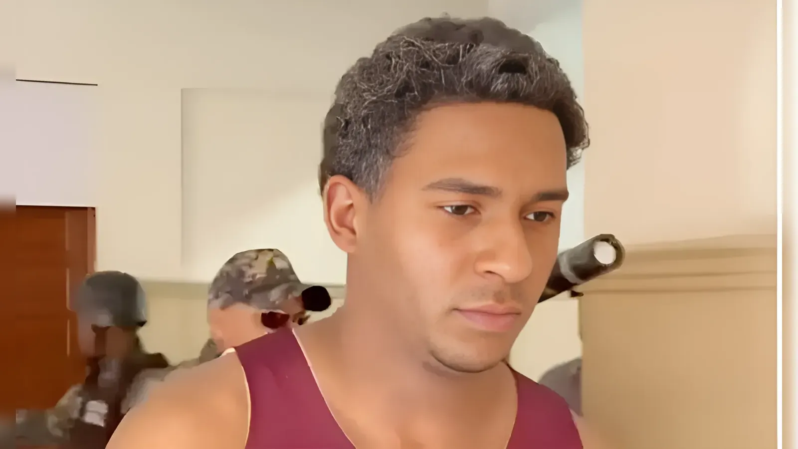Close-up portrait of a man in a maroon sleeveless shirt, looking downward in a room with blurred figures in the background.