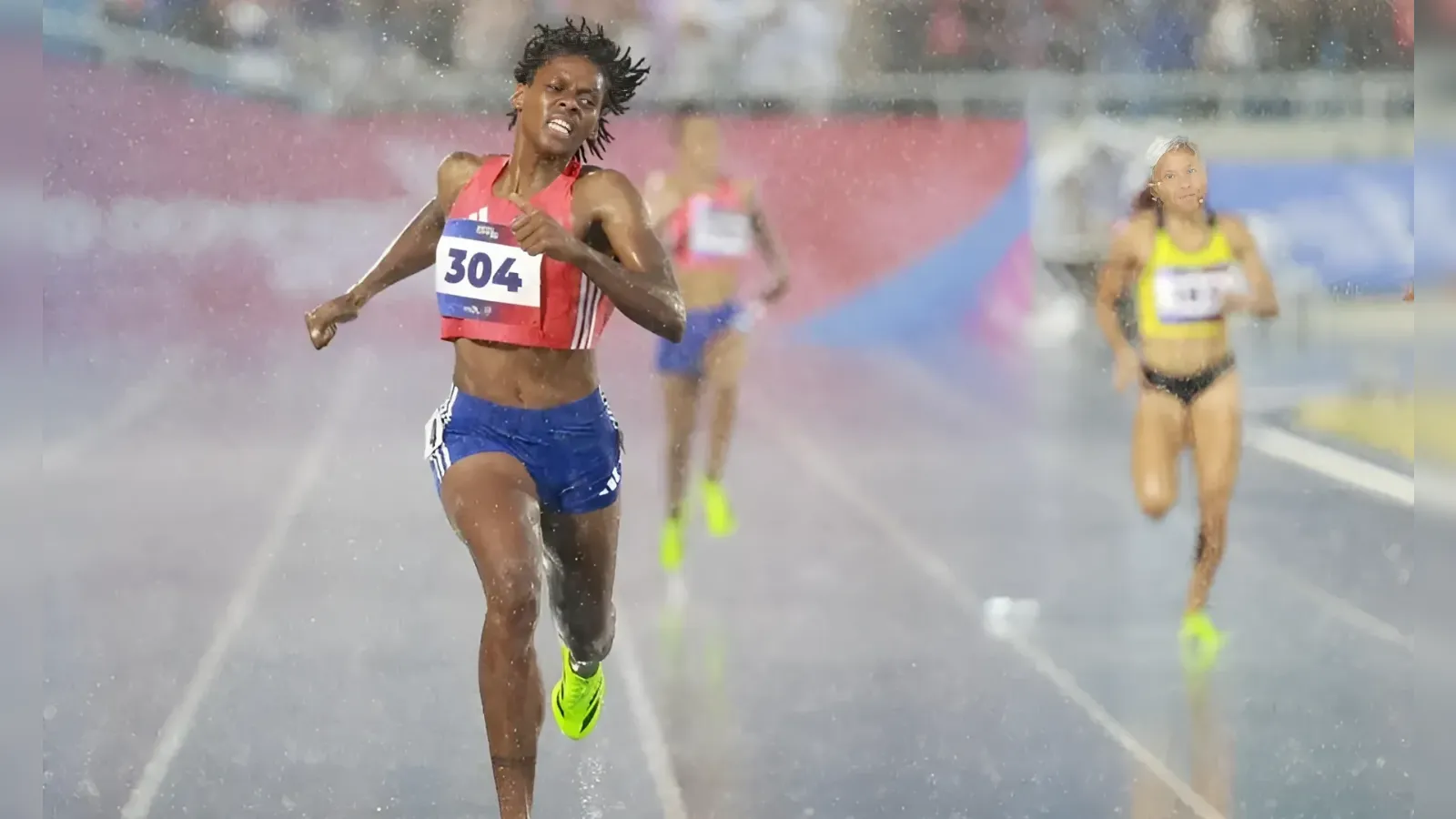 Female sprinter in a red top and blue shorts (bib 304) sprinting on a rainy track.