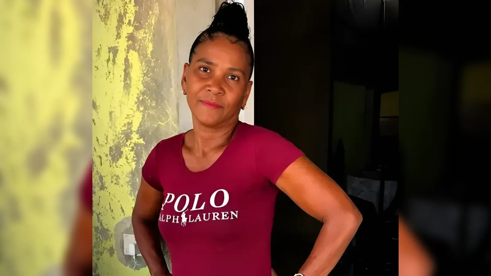 Portrait of a woman with a bun, wearing a maroon Polo Ralph Lauren t-shirt, hands on hips, indoors by a yellow-green wall.