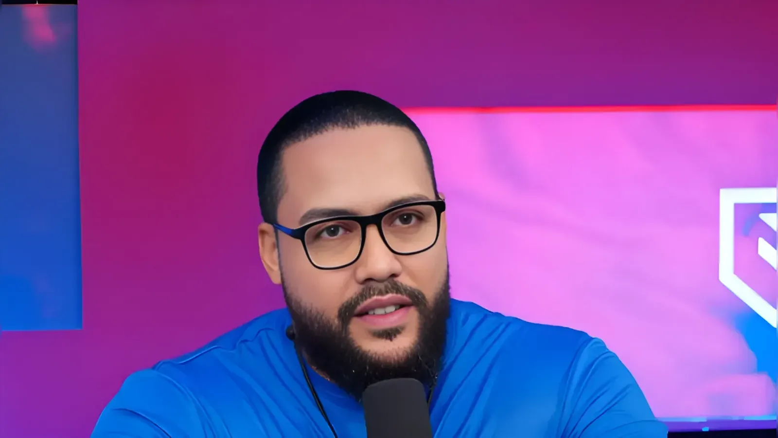 Bearded man wearing black glasses speaks into a microphone in a purple studio setting.