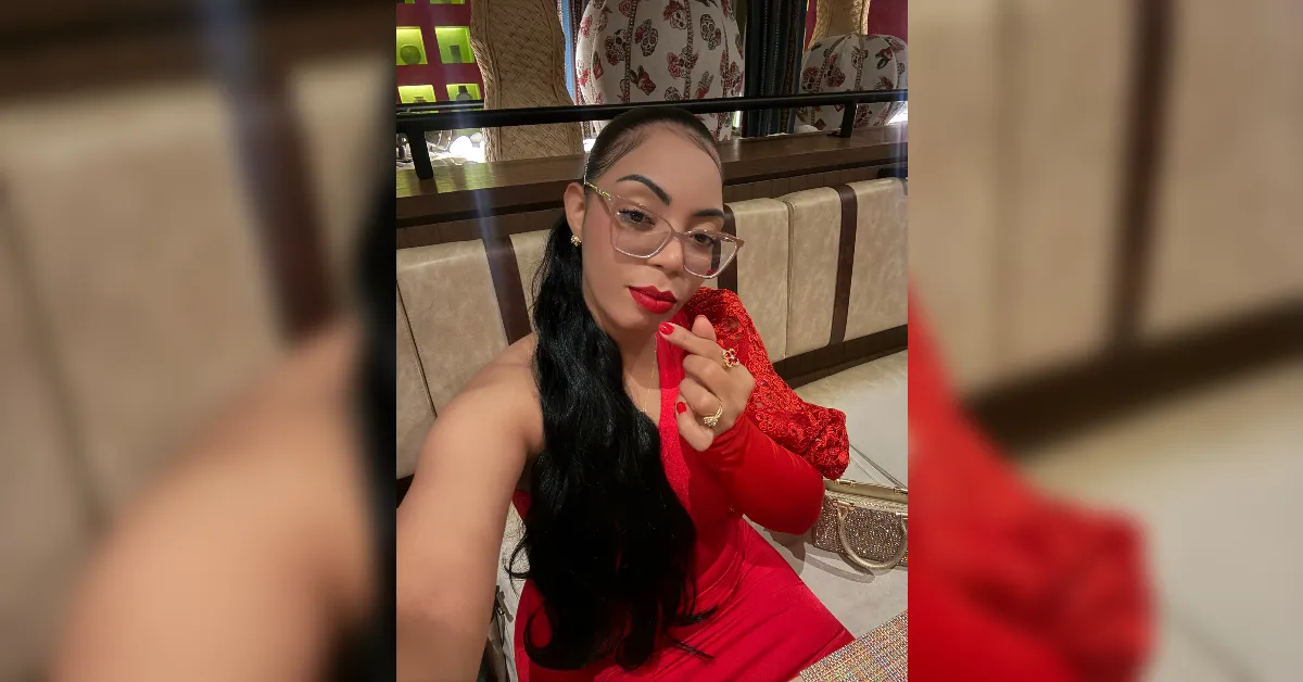Woman in a red dress taking a selfie in a stylish indoor setting, wearing gold-rimmed glasses and bright red lipstick with long black hair cascading over her shoulder.