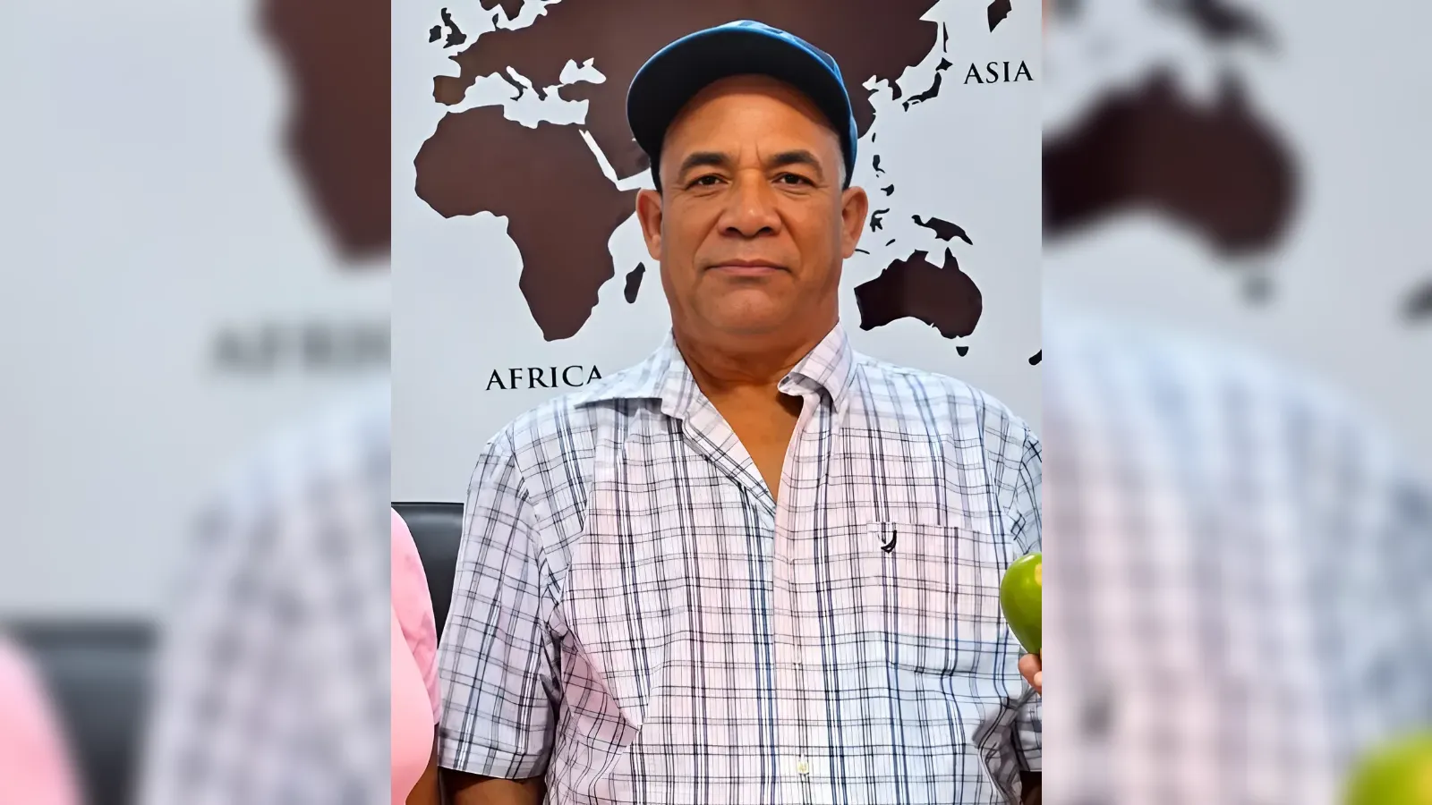 Middle-aged man in a blue cap and plaid shirt, holding a green apple in front of a world map wall backdrop.