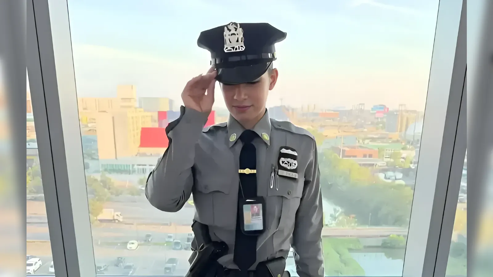Young security guard in a gray uniform salutes while tipping a peaked cap with a badge, standing by a large window with a city view behind him.