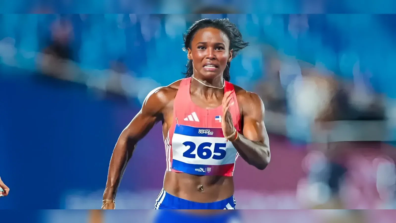 Female sprinter in a red top with bib 265, sprinting on a blue track in a stadium.